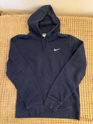 Mörkblå hoodie från Nike - Snygg mörkblå hoodie från Nike med klassisk vit swoosh-logga på bröstet. Hoodien har huva med dragsko och en stor magficka framtill. Tillverkad i mjukt bomullsmaterial som är skönt att ha på sig. Perfekt för en avslappnad och sportig stil.