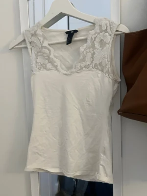 Vit spetsdetaljerad linnetopp H&M - Säljer en vit linnetopp från H&M med v-ringning och breda spetsdetaljer upptill. Toppen är ärmlös och har en mjuk, bekväm passform. Perfekt att styla med jeans eller kjol för en fräsch look.
