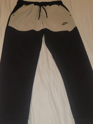 Nike-tech mjukisbyxor - Snygga mjukisbyxor från Nike i svart och beige. Byxorna har dragsko i midjan, två sidofickor och Nike-logga på vänster sida. Tillverkade i mjukt och bekvämt material, perfekta för chill eller träning.