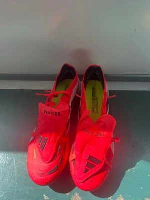 Röda Adidas Predator fotbollsskor elite - Snygga röda Adidas Predator fotbollsskor med svarta detaljer och texten 'EST. 1949' på plösen. Skorna har snörning, låg profil och en yttersula med fasta dobbar för bra grepp på planen. Ovandelen är i syntetmaterial och designen är riktigt sportig.