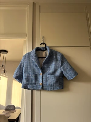 Kort blå tweedkavaj med fickor - Trendig kort kavaj i blå tweed med två stora framfickor och guldknappar. Kavajen har korta ärmar och klassisk krage, perfekt för en cool och stilren look. Materialet är strukturerat och ger en exklusiv känsla.