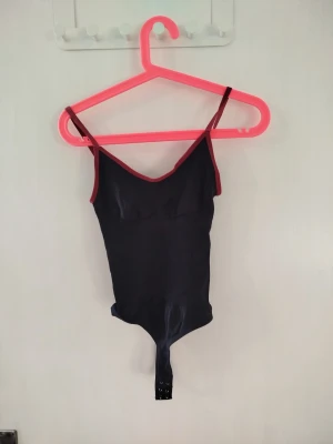 Svart body med vinröda detaljer XS - Snygg svart body med tunna axelband och vinröda kanter. Tillverkad i stretchigt material för en snygg och bekväm passform. Perfekt att styla till en kjol eller till ett par jeans. Vet tyvärr inte vart den kommer ifrån.