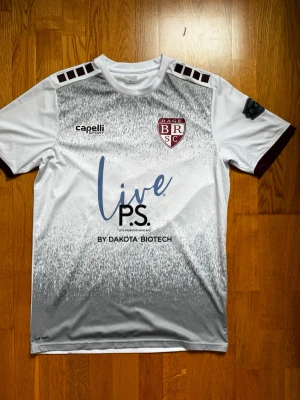 Vit och vinröd fotbollströja Capelli - Snygg fotbollströja från Capelli i vitt med vinröda detaljer och tryck. Tröjan har klubbmärke på bröstet, sponsorloggor och nummer 13 på ryggen. Tillverkad i lätt och ventilerande funktionsmaterial, perfekt för träning eller match.