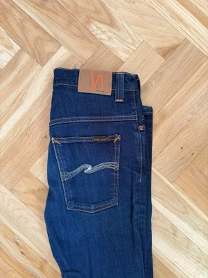 Mörkblå jeans från Nudie Jeans - Säljer ett par mörkblå jeans från Nudie Jeans med klassisk femficksmodell och kontrastsömmar. Jeansen har en rak passform och är tillverkade i mjuk denim med Nudie-logga på bakfickan och läderpatch i midjan.