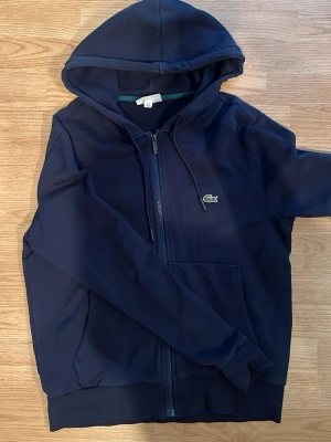 Mörkblå hoodie från Lacoste - Snygg mörkblå hoodie från Lacoste med dragkedja framtill och klassisk krokodillogga på bröstet. Hoodien har huva med dragsko och praktiska fickor på magen. Perfekt för en avslappnad och sportig stil. Materialet känns mjukt och bekvämt.