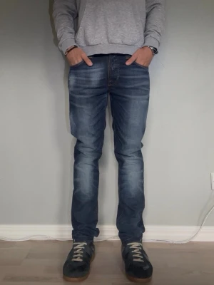 Blå slim fit jeans med slitningar - NUDIE JEANS THIN FINN — BROKEN ORANGE  Storlek: W30 L32 Passform: Slim fit Material: 98% ekologisk bomull, 2% elastan Modell: Thin Finn Tvätt: Broken Orange  Thin Finn är Nudies klassiska slim-modell — sitter tätt längs hela benet utan att vara tight. Broken Orange-tvätten ger ett mörkblått, avskalat uttryck med subtila fading-detaljer som passar året runt..  Modellen på bilden är 175 cm och väger 65.   Skick: Mycket bra skick utan defekter. Nypris: ca 1 800 SEK