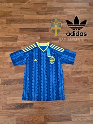 Blå Sverige fotbollströja Adidas - 🟦 Sveriges VM Bortatröja – Adidas – Storlek S 🟦 Officiell Sveriges VM bortatröja från Adidas i blå design med gula detaljer. Klassisk landslagslook med unikt mönster i tyget, gul V-ringning och Adidas tre ränder på axlarna. Svensk landslagsbadge och Adidas-logga på bröstet. Helt ny med tags kvar och tillverkad i ventilerande matchmaterial. Perfekt för samlare eller matchdagar. 📦 Snabb leverans 📩 Kontakta vid frågor eller bilder.