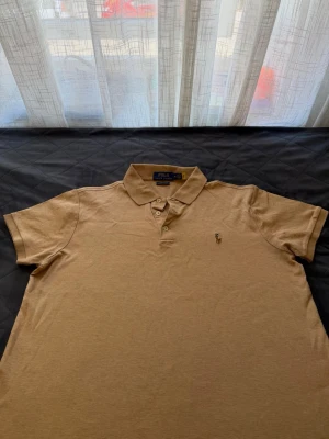 Beige piké från Polo Ralph Lauren - Klassisk beige pikétröja från Polo Ralph Lauren i modellen Custom Slim Fit. Tröjan har korta ärmar, krage och knappar framtill samt den ikoniska broderade loggan på bröstet. Tillverkad i mjuk bomull för en bekväm känsla.
