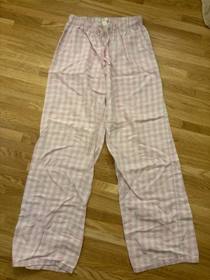 Rutiga pyjamasbyxor från Gina Tricot - Säljer ett par supermysiga pyjamasbyxor från Gina Tricot i ljusrosa och vitt rutigt mönster. Byxorna har resår i midjan och dragsko för extra komfort. Perfekta för chillkvällar hemma. Materialet känns mjukt och skönt mot huden.