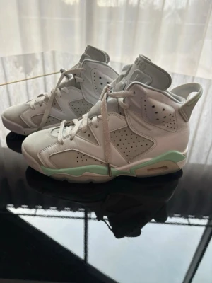 Air Jordan 6 vita och mint sneakers - Air Jordan 6 sneakers i vitt läder och mocka med mintgröna detaljer på sulan. Klassisk hög modell med perforerade paneler, vadderad plös och dragflik bak. Snygga snörhål och ikoniska detaljer som ger en sportig vibe. Perfekta för dig som gillar streetwear och retrostil.
