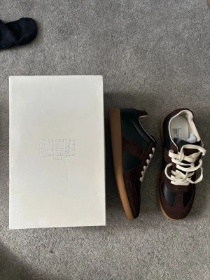 Maison Margiela sneakers brun/grön - Snygga sneakers från Maison Margiela i brunt och mörkgrönt läder med mockadetaljer och vita skosnören. Klassisk låg modell med gummisula och stilren design. Perfekta för dig som vill ha något unikt och trendigt.
