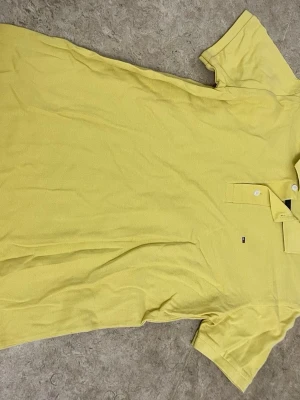 Gul pikétröja från Tommy Hilfiger - Snygg gul pikétröja från Tommy Hilfiger med klassisk krage och två knappar framtill. Tröjan har korta ärmar och den ikoniska Hilfiger-loggan broderad på bröstet. Perfekt för en fräsch och stilren look.