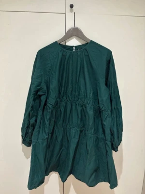 Grön långärmad klänning ASOS DESIGN - Mörkgrön klänning från ASOS DESIGN med lång ärm och rund halsringning. Klänningen har en lös passform och är sydd i flera sektioner för en volangliknande effekt. Perfekt för dig som gillar enkel men snygg stil.