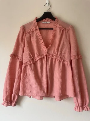 Rosa volangblus från NA-KD - Supersöt rosa blus från NA-KD med volangdetaljer längs ringningen och bröstet. Blusen har en djup v-ringning, långärmade puffärmar med resår vid ärmslut och är gjord i ett lätt, strukturerat tyg som känns luftigt och bekvämt.