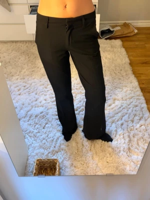 Svarta utsvängda kostymbyxor - Snygga svarta kostymbyxor med utsvängda ben och hällor för skärp. Klassisk midja och stilren design som passar perfekt till en trendig look. Materialet ser ut att vara en mjuk och följsam mix, perfekt för dig som gillar stilrena och bekväma plagg. Som nya!