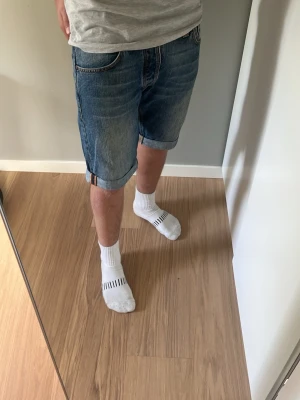 Blå nudie jeansshorts - Klassiska blå jeansshorts från Nudie med upprullad kant och femficksmodell. Shorts i normal passform med midja och slitna detaljer på bakfickorna. Perfekta för en avslappnad stil under varmare dagar.