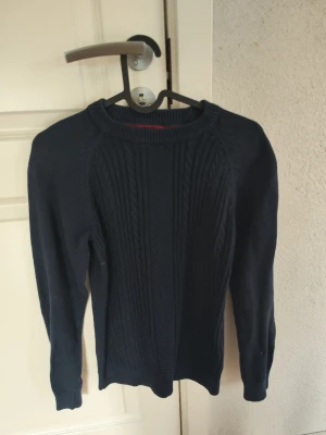 Mörkblå kabelstickad tröja från Hampton republic - Säljer en mörkblå kabelstickad tröja med rund hals och långa ärmar. Tröjan har ett klassiskt mönster framtill och ribbade muddar vid ärmslut och nederkant. Perfekt för dig som gillar enkel men snygg stil. Passar de runt 13 år. Knappt använd, nästan helt ny.