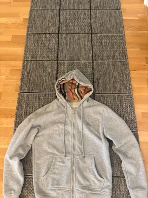 Grå zip hoodie från Burberry - Snygg grå zip hoodie från Burberry med klassiskt rutigt foder i huvan och diskret broderad logga på bröstet. Hoodien har dragsko i huvan, två fickor fram och är tillverkad i mjukt bomullsmaterial. Perfekt för en avslappnad och stilren look.