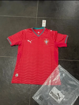 Portugal röd fotbollströja Puma - Snygg röd Portugal fotbollströja från Puma med vita vågmönster och gröna detaljer vid krage och ärmslut. Tröjan har korta ärmar, V-ringning och Portugal-emblem på bröstet. Perfekt för match eller träning.