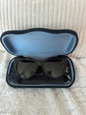 Solglasögon Gucci - Äkta svarta/skölpadsfärgade solglasögon från Gucci med breda bågar och mörka glas. Klassisk cat-eye form och diskret Gucci-logga på sidorna. Kommer i originalfodral med putsduk och dustbag. Inga synliga tecken på användning förutom på insidan av bågarna. 