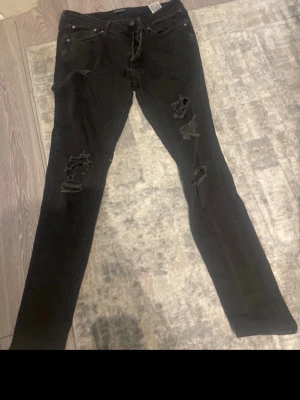 Svarta ripped skinny jeans - Säljer ett par svarta skinny jeans med slitningar och hål på benen för en edgy look. Jeansen har klassisk femficksmodell och dragkedja med knapp. Perfekta för dig som gillar streetstyle och vill ha en tuff vibe i garderoben.