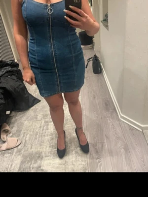 Blå jeansklänning med dragkedja - Snygg blå jeansklänning med smala axelband och markerad söm framtill. Klänningen har en hel dragkedja med ringdetalj framtill och är kort i modellen. Perfekt för dig som gillar en tight passform och vill ha en enkel men trendig look.