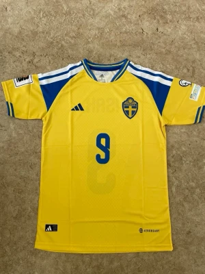 Sverige matchtröja Isak #9 Adidas - Gul och blå Sverige matchtröja från Adidas med Isak och nummer 9 på ryggen. Tröjan har korta ärmar, rund hals och detaljerade tryck på ärmarna. Tillverkad i lätt och ventilerande material (AEROREADY), perfekt för fotbollsträning eller match.