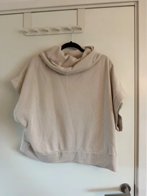 Beige hoodie med korta ärmar - Säljer en beige hoodie med korta ärmar och bred passform. Hoodien har en stor huva och är gjord i mjukt sweatshirtmaterial, perfekt för en avslappnad och trendig look. Inga synliga loggor eller tryck. Sparsamt använd. Perfekt för oversized look.