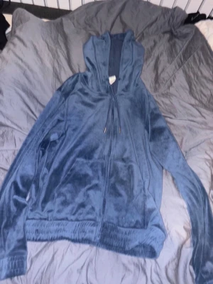 Blå velour hoodie med dragkedja - Mjuk och skön hoodie i blå velour med dragkedja framtill och stora fickor. Tröjan har huva med snören och ribbade muddar vid ärmslut och nederkant. Perfekt för en chill och avslappnad stil.