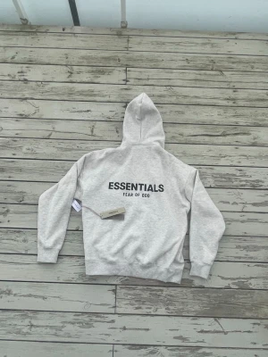Grå Essentials Fear of God hoodie - Snygg ljusgrå hoodie från Essentials Fear of God med svart logga tryckt både på bröstet och ryggen. Klassisk passform med huva och ribbade muddar. Perfekt för en avslappnad och trendig streetwear-look. Materialet känns mjukt och bekvämt.