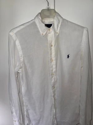 Vit linneskjorta från Ralph Lauren - Klassisk vit skjorta i linne från Ralph Lauren med lång ärm och knappar framtill. Skjortan har button-down krage och den ikoniska mörkblå loggan broderad på bröstet. Perfekt för en clean och stilren look. Slimfit