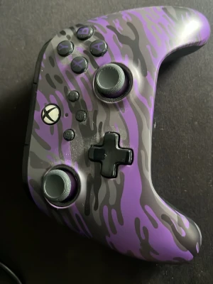 PowerA Xbox handkontroll lila camo - Säljer en PowerA handkontroll till Xbox med snyggt lila och grått camo-mönster. Kontrollern har svarta detaljer, texturerade grepp och extra knappar på baksidan. Perfekt för gaming och har en cool design som sticker ut bland andra kontroller.