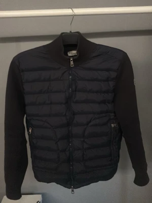 Svart cardigan från Moncler - Snygg svart cardigan från Moncler med quiltad front och mjuka stickade ärmar. Jackan har hög krage, två dragkedjefickor och Moncler-logga på ärmen. Perfekt för kalla dagar och ger en clean look. 