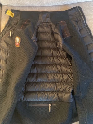 Svart dunväst från Parajumpers - Snygg svart dunväst från Parajumpers med quiltad framsida, meshpaneler på sidorna och ikonisk PJS-patch på axeln. Västen har dragkedja, orange detalj vid kragen och praktisk ficka med dragkedja. Perfekt för lager-på-lager och streetwear.