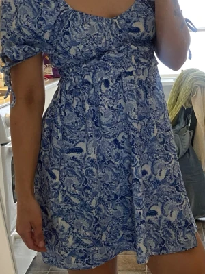 Blåvit kortklänning med puffärm - Söt kortklänning i blått och vitt med paisley-inspirerat mönster. Klänningen har puffärmar med knytband och en smickrande, lätt utställd passform. Perfekt för varma dagar och har en luftig känsla. 