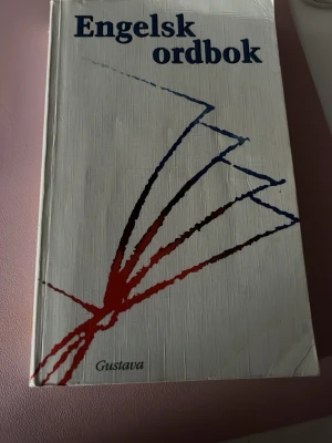 Engelsk ordbok från Gustava - Engelsk ordbok med cirka 75 000 ord, uttryck och fraser. Boken har vit hårdpärm med abstrakt röd och blå design på omslaget. Perfekt för både skolan och vardagsbruk när du vill slå upp engelska ord snabbt och enkelt.