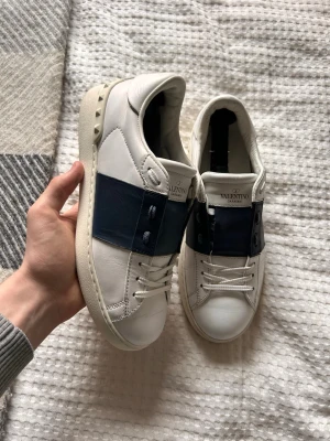 Valentino vita sneakers med blå detalj - Säljer ett par vita sneakers från Valentino med bred mörkblå läderdetalj över sidan och klassiska nitar på hälen. Skorna har vita snören och en chunky sula. Perfekta för dig som gillar stilrena men ändå unika sneakers.