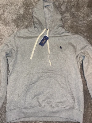 Grå hoodie från Polo Ralph Lauren - Snygg grå hoodie från Polo Ralph Lauren med klassisk huva och vita dragsnören. Har en känguruficka framtill och den ikoniska lilla loggan broderad på bröstet. Tillverkad i mjuk bomullsblandning som känns skön mot huden. (Rep)