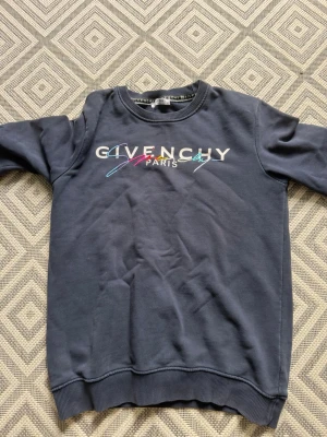 Mörkblå sweatshirt från Givenchy - Säljer en mörkblå sweatshirt från Givenchy med färgglad logotyp och texten 'Paris' på bröstet. Tröjan har rund halsringning, långa ärmar och ribbade muddar vid ärmslut och nederkant. Perfekt för en trendig och avslappnad stil.