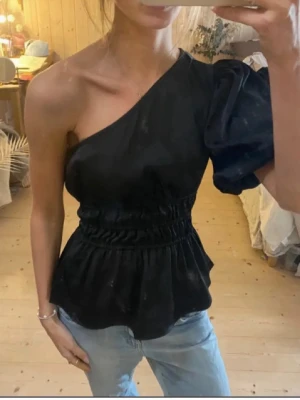 Svart one shoulder topp med puffärm - Snygg svart topp med one shoulder-design och en voluminös puffärm. Midjan är markerad med smockdetaljer och nederdelen har en lätt utsvängd peplumform. Perfekt för dig som vill sticka ut lite extra. Materialet ser ut att vara mjukt och glansigt.
