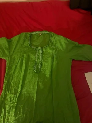 Grön klänning med knappar och veck - Grön klänning i lätt material med rundad halsringning, dekorativa knappar framtill och veckdetaljer vid bröstet. Lång ärm och loose passform för en chill vibe. Perfekt för dig som gillar färg och enkel design.