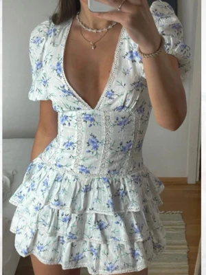 Ny  populär Vit blommig kortklänning med volanger - Supersöt vit kortklänning med blå och lila blommor, puffärmar och djup v-ringning. Klänningen har flera lager volanger i kjolen och spetsdetaljer längs sömmarna. Perfekt för sommarens alla tillfällen.