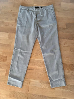 Ljusgrå kostymbyxor med pressveck - Snygga ljusgrå kostymbyxor med raka ben och pressveck framtill. Byxorna har bälteshällor, sidofickor och två bakfickor med knapp. Materialet ser ut att vara en tunnare, vävd kvalitet som passar till en dressad look.