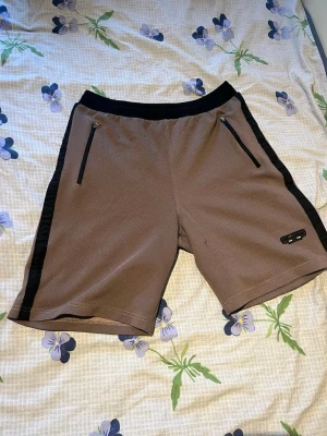 Bruna shorts med svarta detaljer - Säljer ett par bruna shorts med svarta sidor och midja. Shortsen har dragkedjeförsedda fickor framtill och en liten logga på vänster ben. Perfekta för en avslappnad stil.