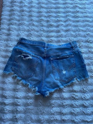 Blå jeansshorts med fransig kant - Säljer ett par blå jeansshorts med rå, fransig kant och slitningar på bakfickorna. Shortsen har två bakfickor, bälteshällor och klassisk femficksdesign. Perfekta för en avslappnad och trendig sommarstil.