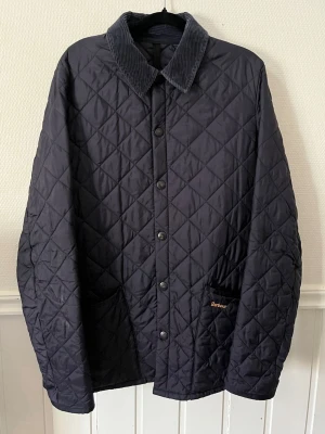 Marinblå quiltad jacka från Barbour - Nypris 2200kr. Storlek S.Klassisk marinblå quiltad jacka från Barbour med knäppning framtill och två fickor. Jackan har en mjuk manchesterkrage och diskret Barbour-logga på fickan. Perfekt för höst och vår, tidlös design och bekväm passform.