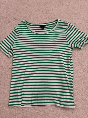 Grönvit randig t-shirt från Monki - Säljer en grön och vit randig t-shirt från Monki. T-shirten har rund halsringning och korta ärmar. Tillverkad i mjuk bomull som känns skön mot huden. Perfekt för dig som gillar en enkel men snygg stil.