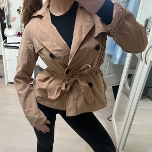 trenchcoat - Snygg beige trenchcoat med knytbälte och svarta knappar. Perfekt för vår och höst. Jackan har långa ärmar och en klassisk krage.