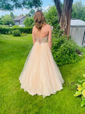 Champagnefärgad balklänning med snörning - Elegant långklänning i champagnefärg med blänkande stenar på överdelen och snörning i ryggen. Klänningen är axelbandslös och har en lös underdel till som gör att klänningen blir fluffigare (följer med vid köp). Perfekt för bal eller liknande! Storlek:S/M & i kuppa 75/80 B-C (Inga defekter på förutom at några stenar sitter löst men jätte lätt att sy fast hårdare) går att hämta i Norrköping 