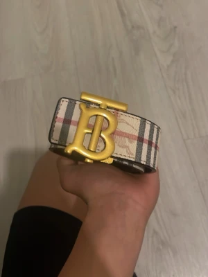 Randigt Burberry bälte med guldspänne - Snyggt bälte från Burberry med klassiskt beige, svart och rött rutmönster och ett stort guldigt B-spänne framtill. Perfekt accessoar för att lyfta vilken outfit som helst. Tillverkat i skinn med exklusiv känsla och ikonisk design.